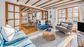 A spacious living room with a blue sofa, a grey sofa, and a wicker ottoman. There is a television on a wooden stand and a large window with curtains. - Charming Family Stables Conversion - Pass the Keys (Emsworth)