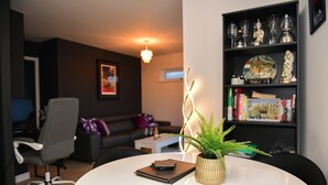 Un salon moderne avec une table ronde blanche, un fauteuil de bureau gris, un canapé noir, une petite table d’appoint blanche, une plante en pot et une bibliothèque agrémentée d’objets décoratifs.