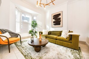 Ein Wohnzimmer mit einem grünen Sofa, einem orangefarbenen Sessel und einem runden Couchtisch mit Flasche und Gläsern.