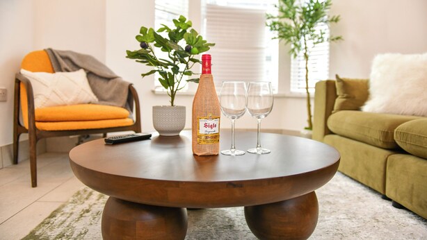Een ronde houten salontafel met een fles, twee glaasjes, een afstandsbediening en een bloempot.