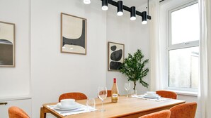 Een eethoek met een houten tafel, oranje stoelen, witte borden, wijnglazen en een fles wijn. Op de muren hangen abstracte schilderijen en op de tafel staat een potplant.