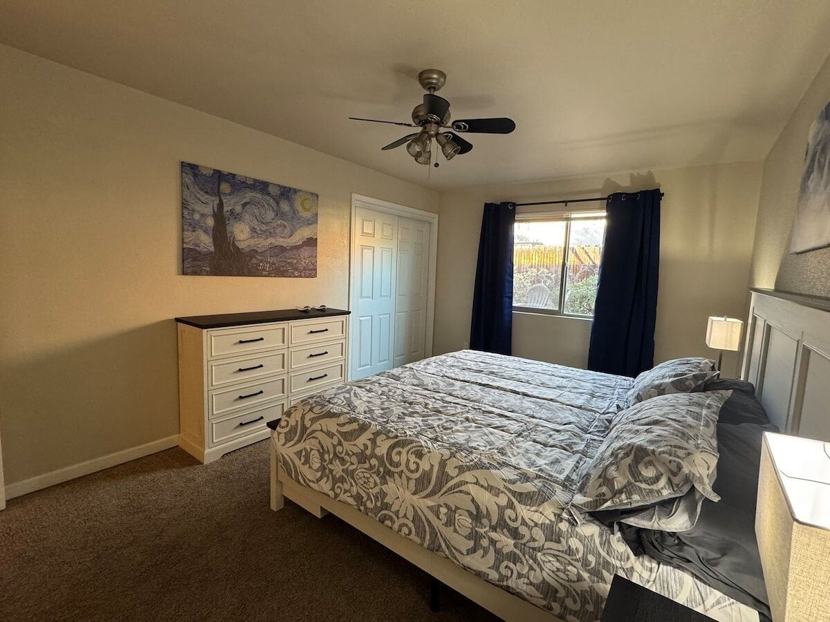 Une chambre à coucher comprenant un lit, une commode, un ventilateur de plafond, une porte et une fenêtre avec des rideaux.