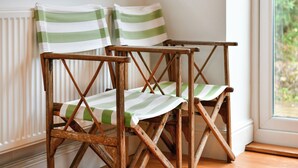 Deux chaises en bois, dotées de coussins à rayures vertes et blanches, sont placées près d’une fenêtre donnant sur un paysage verdoyant.