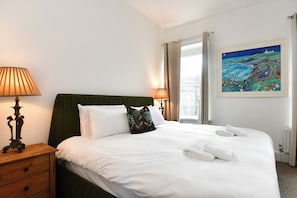 Ein ordentlich bezogenes Bett mit weißen Leinentüchern und zwei Kissen, ein Nachttisch mit einer Lampe und ein Bild an der Wand.