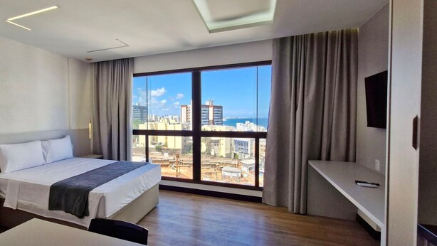 Quarto de hotel com cama, escrivaninha, televisão e uma grande janela com vista para a cidade.