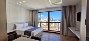 A hotel room with a bed, a desk, a TV, and a large window with a city view. - COARM0111 - Condominio LIT 760 (Salvador)
