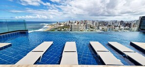 Ein Infinity-Pool mit Liegestühlen, Blick auf die Stadt und ein Strand im Hintergrund.