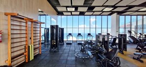 Ein Fitnessraum mit Trainingsgeräten, einer Klimmzugstange und großen Fenstern mit Blick auf ein Gewässer.