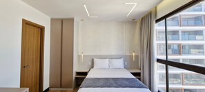 Ein Hotelzimmer mit Bett, Nachttischen, einem Kleiderschrank und einem großen Fenster mit Blick auf Gebäude.