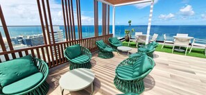 Een balkon met rieten stoelen en tafels, uitzicht op zee en een stadsachtergrond.