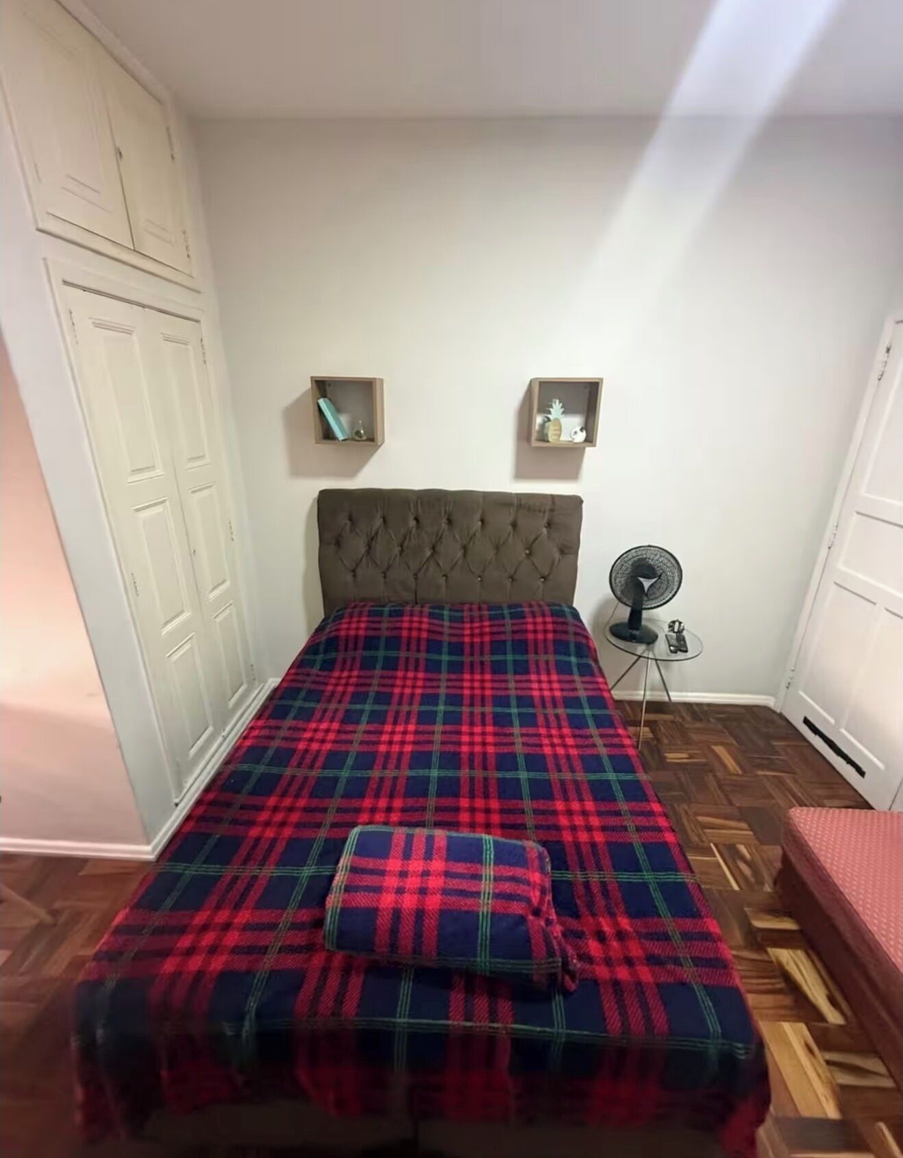 A bedroom with a bed, a wardrobe, a small table with a fan, and a red bench.