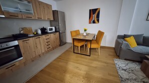 A modern kitchen with wooden cabinets, a microwave, and a dining table with chairs. There's a gray sofa with a yellow pillow and a marble-patterned rug.