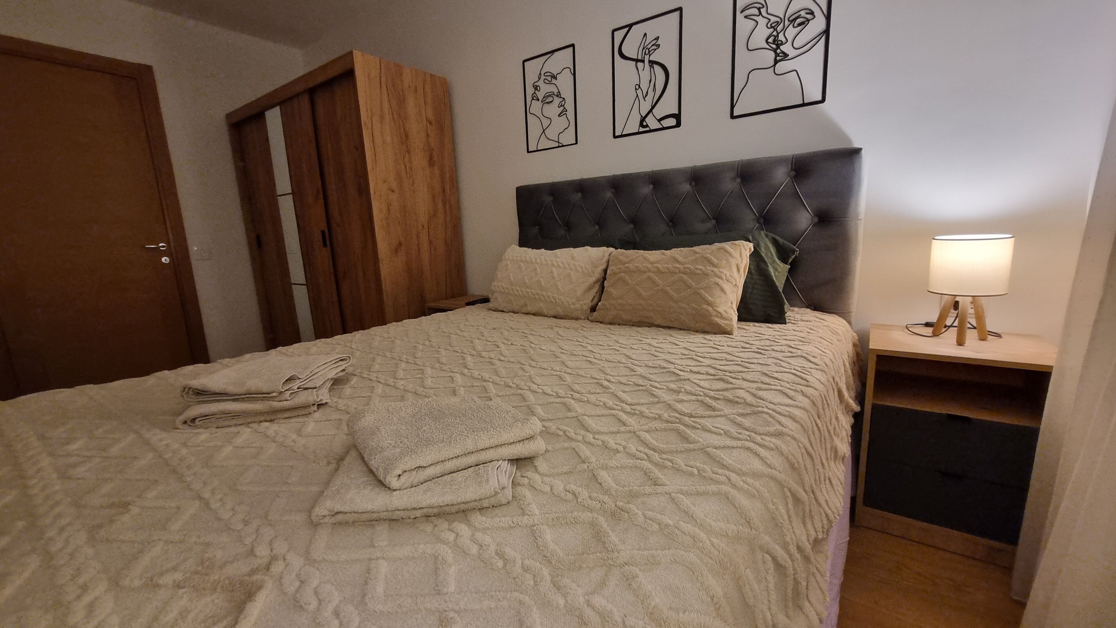 A neatly made bed with a quilted cover, a bedside table with a lamp, and a wooden wardrobe.