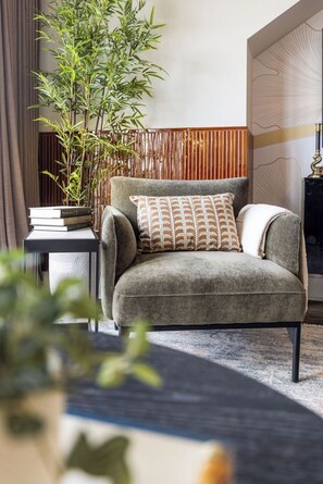 A modern living room with a grey armchair, a patterned throw pillow, a tall bamboo plant, and a small side table with books. - The Lantern Loft | Luxury Riverfront Downtown (Wilmington)