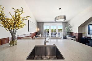 Ett modernt kök med en central köksö, en matplats med soffa och utsikt över omgivningarna genom stora fönster.