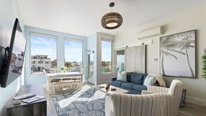Un salon moderne comprenant un canapé, des fauteuils, une table basse et un téléviseur. De grandes fenêtres offrent une vue sur l’extérieur.