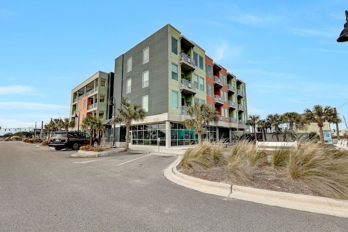 A multi-story building with balconies, a parking lot, and palm trees.