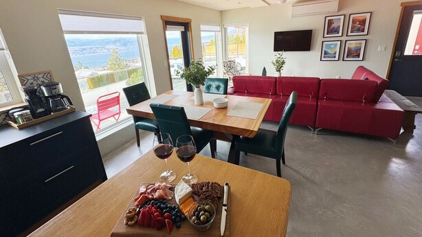 A modern living room with a dining area, a red sofa, and a table set with food and drinks. - Stone & Vines | 2BR Wine Country Retreat | Adventure by Day, Wine by Night (Naramata)