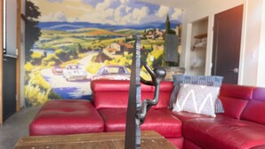 A living room with a red leather sofa, a wooden coffee table, and a wall mural of a countryside road with cars. - Stone & Vines | 2BR Wine Country Retreat | Adventure by Day, Wine by Night (Naramata)