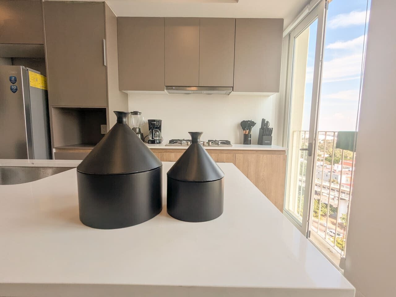 A modern kitchen with two black kitchen scales on the counter, a stainless steel refrigerator, and a window with a city view.