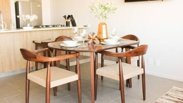 Un comedor con una mesa redonda, cuatro sillas de madera con asientos de mimbre, un florero con flores y una cocina al fondo.