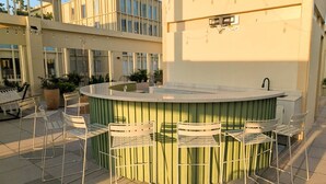 Un espace bar extérieur doté d’un comptoir circulaire, de quatre chaises en métal blanc et d’une rambarde en métal vert.