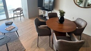 Ett modernt vardagsrum med ett runt matbord i trä, sex stolar, en platt-TV, en stor spegel och utsikt över utomhusmiljön genom ett stort fönster.