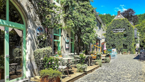 Ruas de paralelepípedos ladeadas por edifícios, com mesas ao ar livre e vegetação.