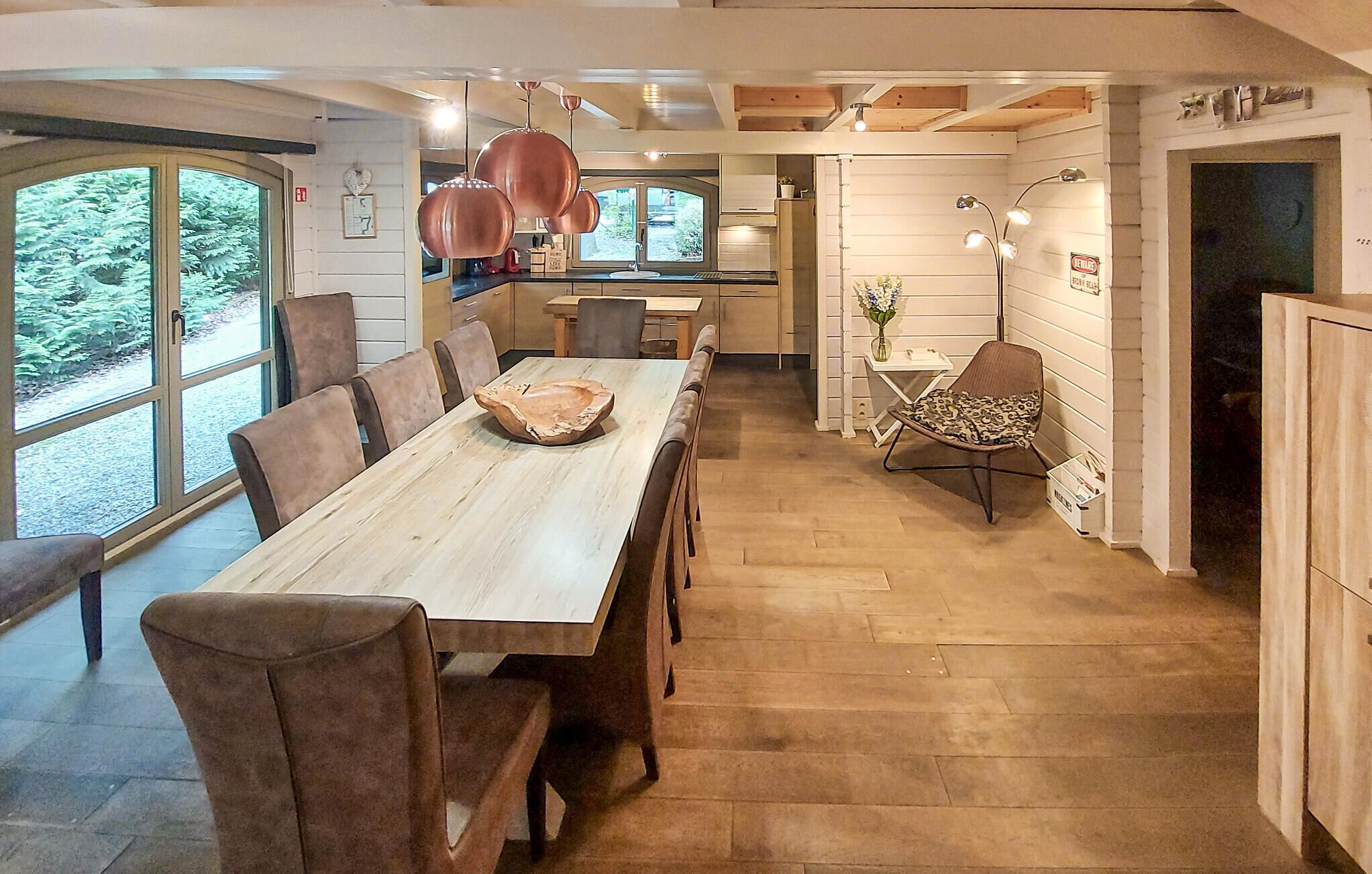 A spacious dining area with a long wooden table and chairs, a kitchen with white cabinets, and a view of greenery through large windows.