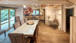 Uma sala de jantar ampla com uma longa mesa de madeira e cadeiras, uma cozinha com armários brancos e vista para o verde através de grandes janelas.