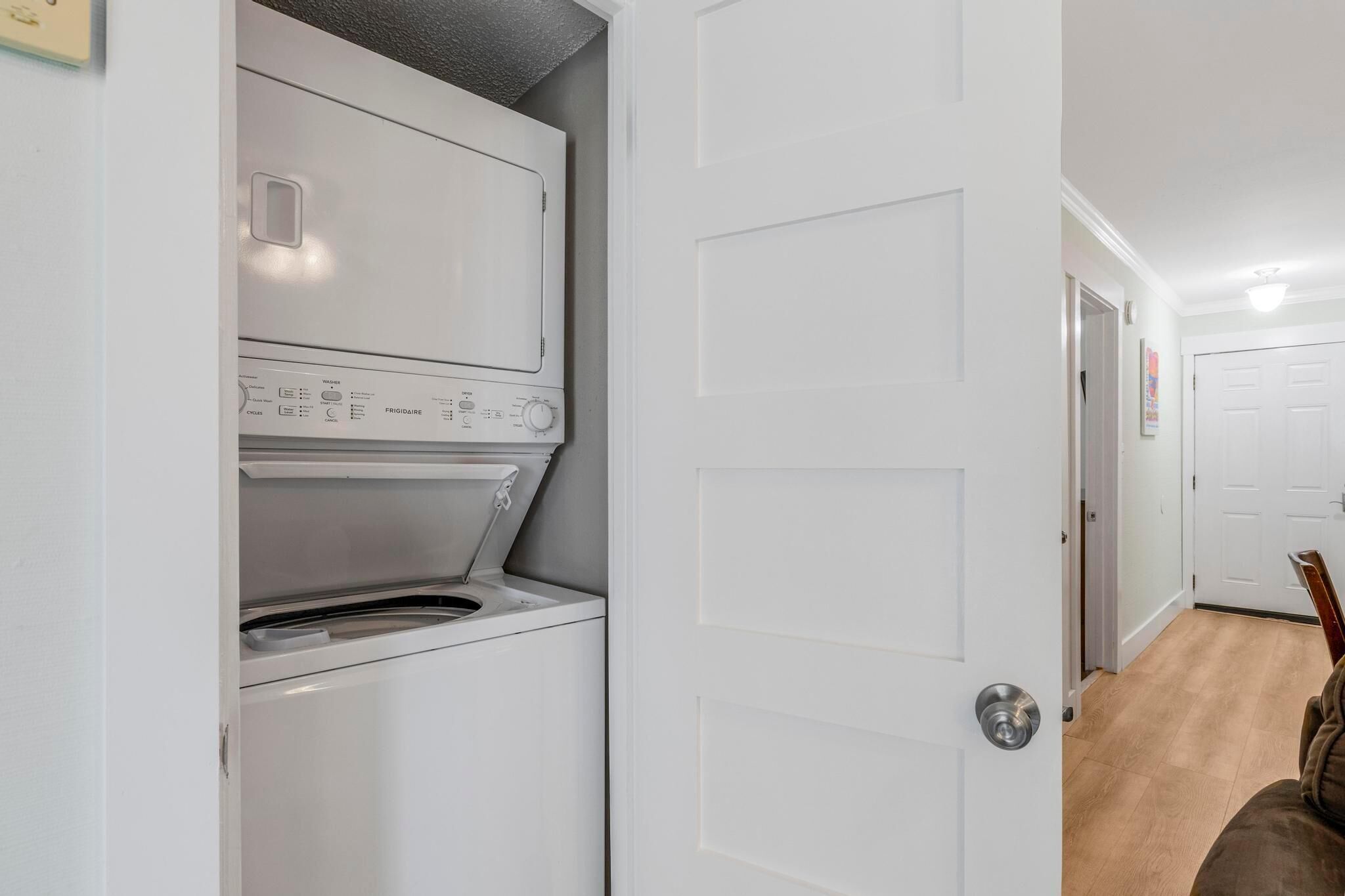 A laundry room with a front-loading washer and dryer, a white door, and a hallway leading to another room.