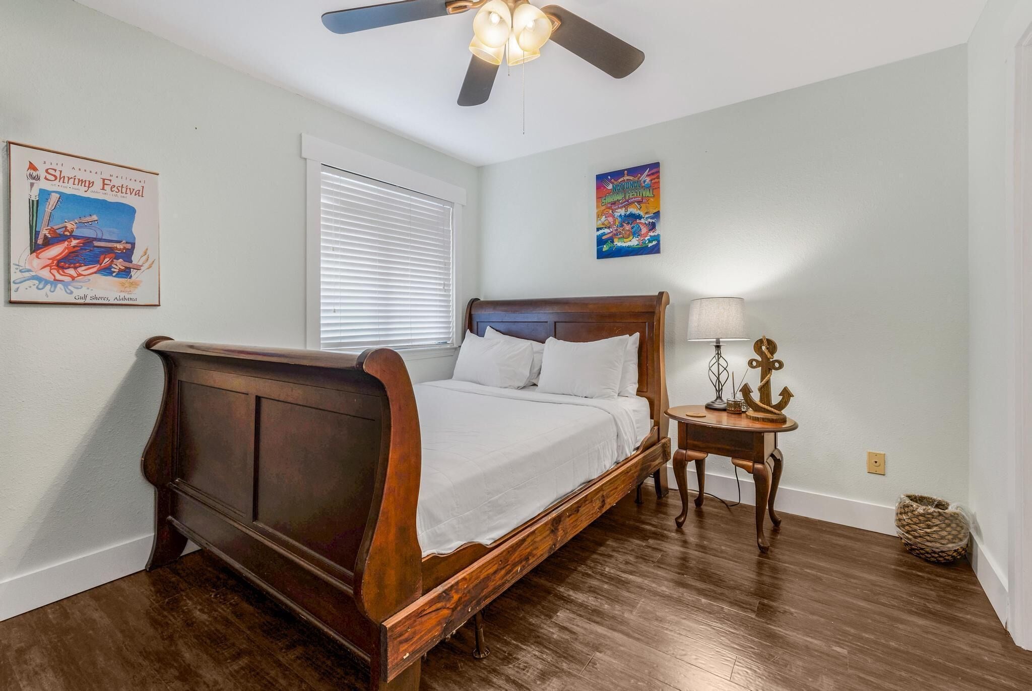 A bedroom with a wooden bed, a ceiling fan, a window with blinds, a small table with a lamp, and wall decorations.