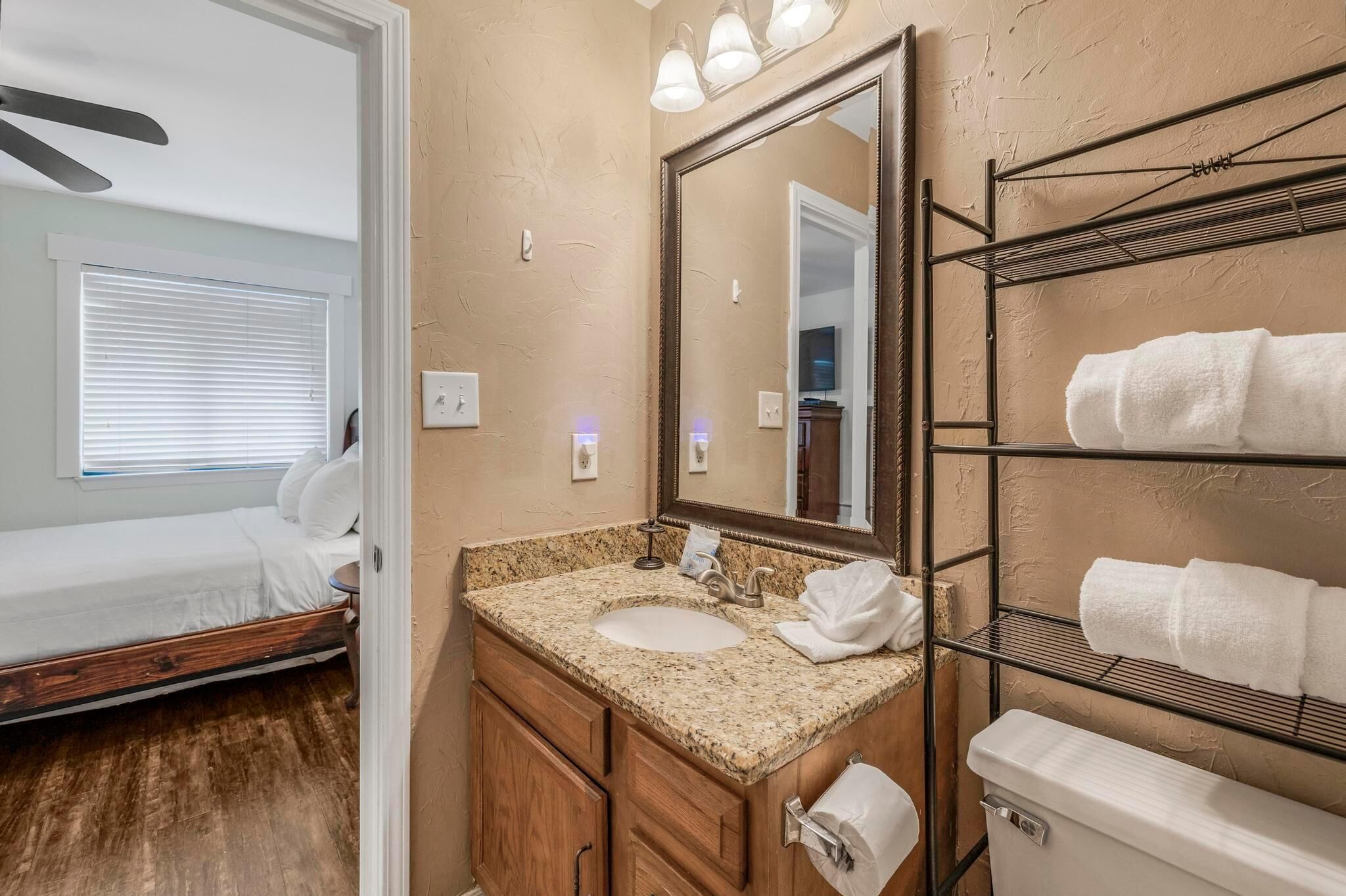 A compact bedroom with a single bed, a bathroom with a sink, mirror, towel rack, and a ceiling fan.