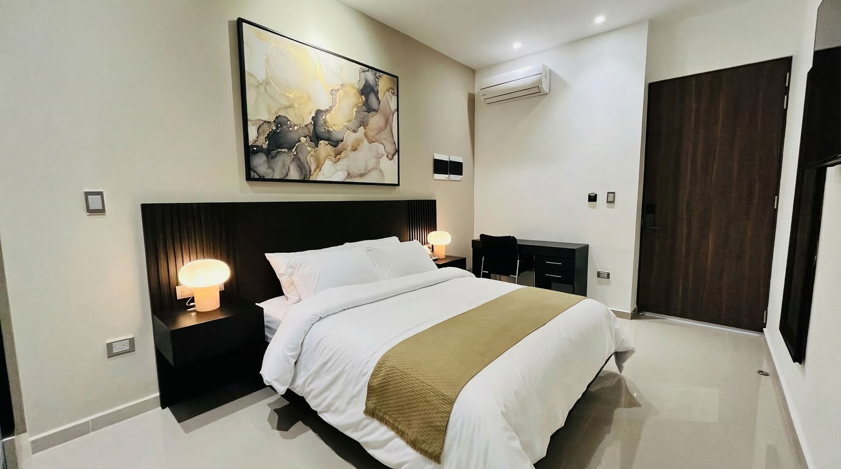 Une chambre d’hôtel moderne avec un grand lit, des lampes de chevet, un tableau, un appareil de climatisation et une porte en bois foncé.