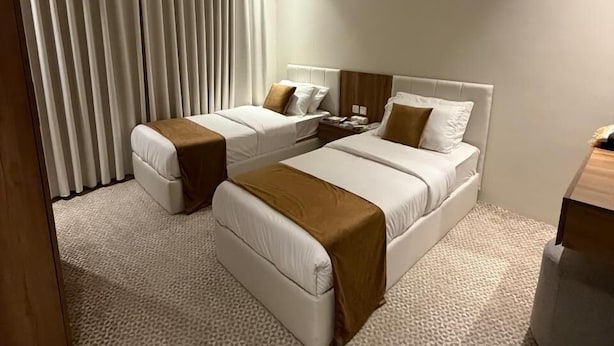 Quarto de hotel moderno com duas camas, uma janela grande com persianas, ar condicionado fixado na parede e um tapete texturizado.