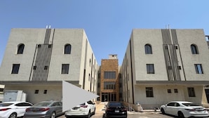 Um edifício moderno com entrada central, duas alas simétricas e área de estacionamento com vários carros.