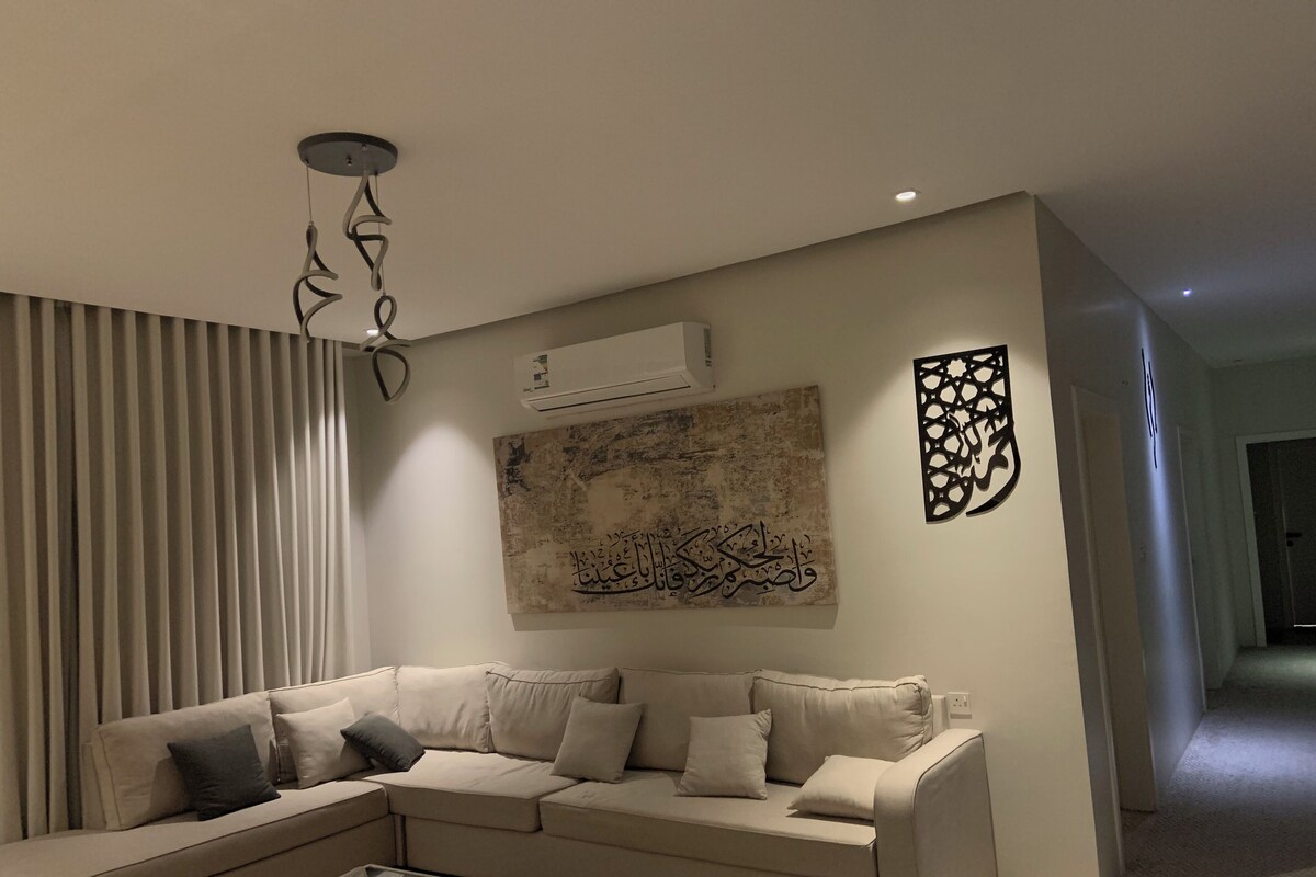 Ein modernes Wohnzimmer mit einem beigefarbenen Ecksofa, einem Glastisch und Wandkunst, darunter arabische Kalligraphie.