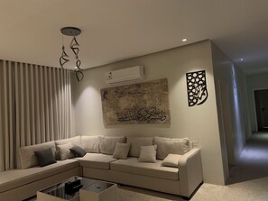 Ein modernes Wohnzimmer mit einem beigefarbenen Ecksofa, einem Glastisch und Wandkunst, darunter arabische Kalligraphie.