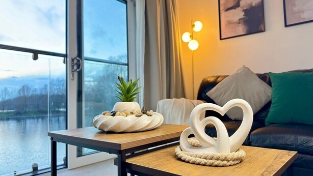 Een moderne woonkamer met uitzicht op een meer, een houten tafel met decoratieve objecten en een bank met kussens.