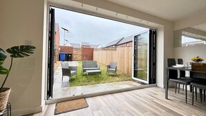 Een moderne woonkamer met een schuifdeur die uitkomt op een achtertuin. De kamer heeft een houten vloer, een eettafel met stoelen en een grote potplant.