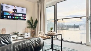 Een moderne woonkamer met een grote televisie waarop een nieuwskanaal wordt vertoond, een schuifdeur naar een balkon met uitzicht op een waterpartij en bomen, en een gezellige zithoek met een leren bank en decoratieve objecten.