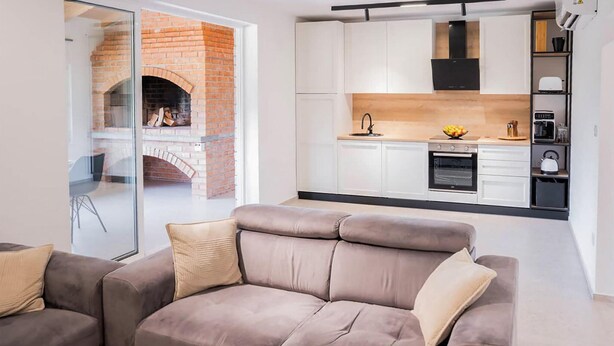 Un salon moderne avec un canapé gris et une porte vitrée donnant sur une cuisine dotée de meubles blancs et d’une cheminée en briques.