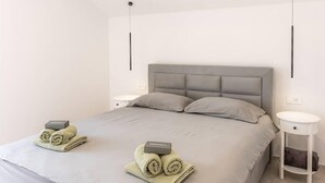 Un lit bien fait, avec une tête de lit grise et deux oreillers, une petite table de chevet avec une lampe, et une serviette enroulée sur laquelle est posé un panneau indiquant « Bienvenue ».