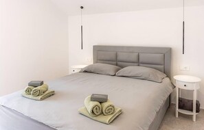 Ein ordentlich gemachtes Bett mit grauem Kopfteil und zwei Kissen, ein kleiner Nachttisch mit Lampe sowie ein zusammengerolltes Handtuch mit der Aufschrift 'Welcome'.