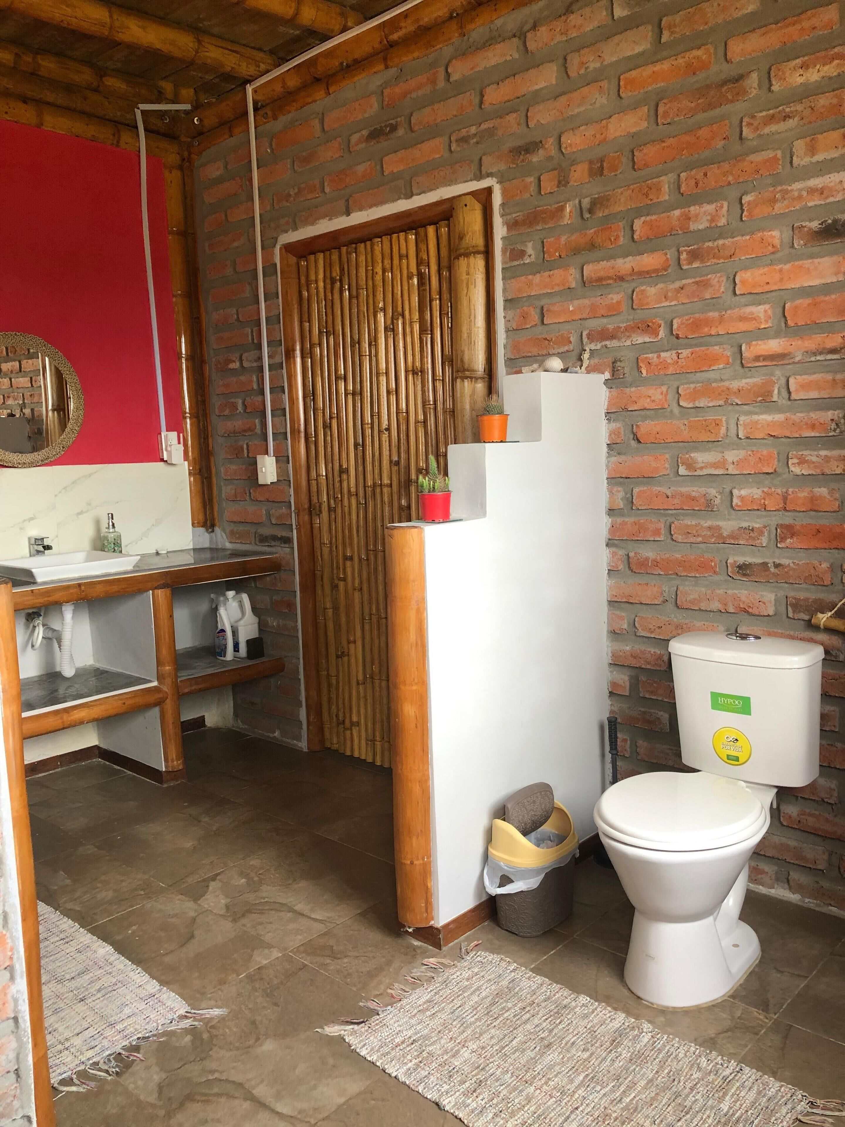 Ein kleines Badezimmer mit Toilette, Waschbecken und Spiegel. Die Wände sind aus freiliegenden Ziegelsteinen und es gibt eine Bambus-Schiebetür.