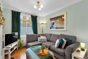 Ein Wohnzimmer mit grauem Sofa, einem Flachbildfernseher, einer Stehlampe und einem Bild an der Wand.