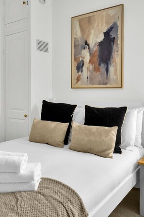 Ein Schlafzimmer mit einem Bett, Kissen, einem an der Wand hängenden abstrakten Gemälde, einer Tür und einem Lüftungsgitter.