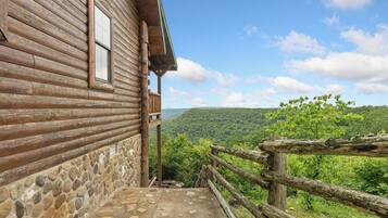 A wooden cabin with a stone foundation, a balcony, and a scenic view of a forested valley.
