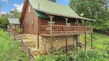 A rustic log cabin with a stone foundation, a wooden porch, and a green metal roof.