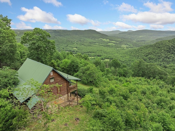 A rustic cabin with a green roof, surrounded by lush greenery and hills in the background. - Rustic Cedar Roost • Private Balcony • Best Views (Jasper)